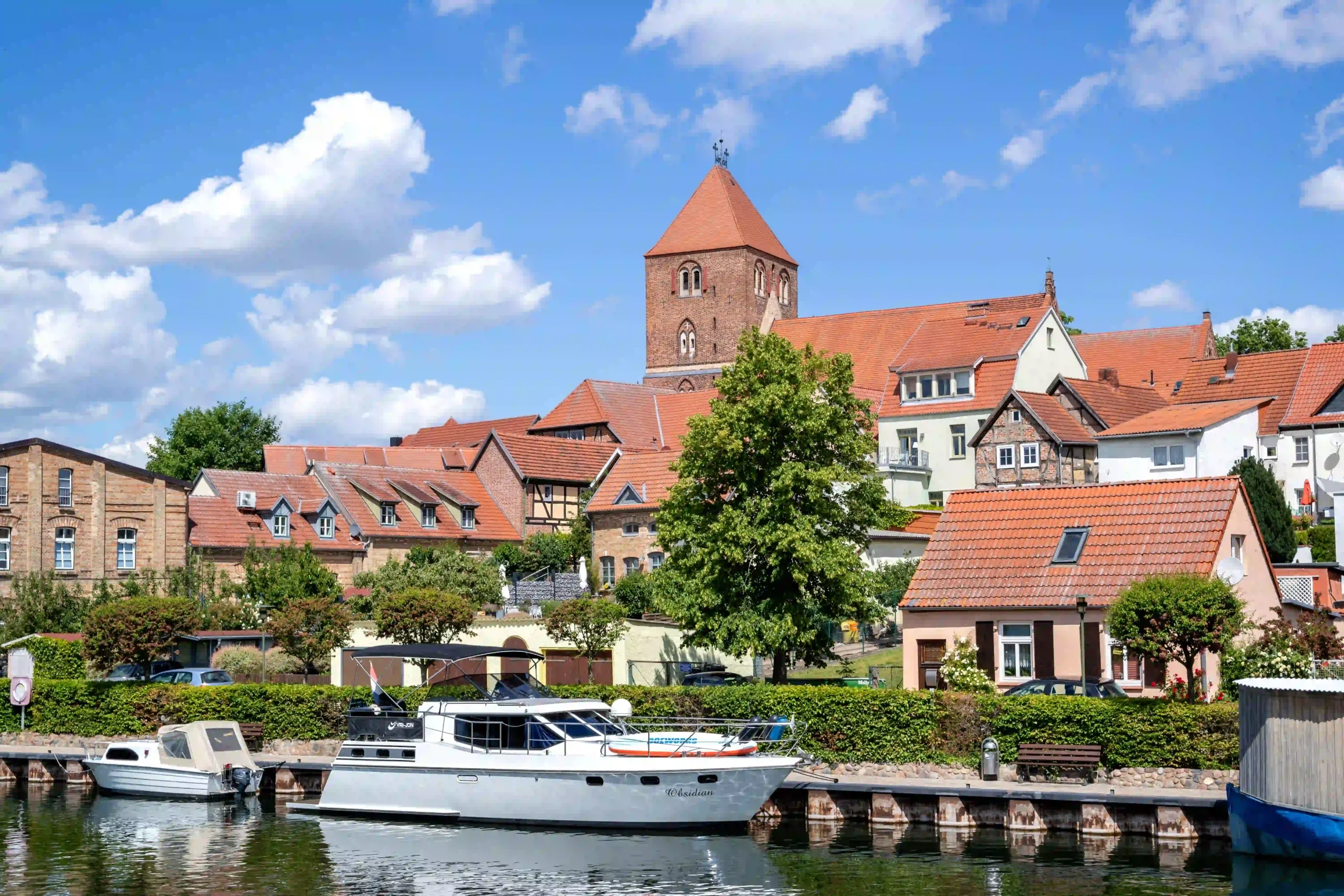Blick auf die Altstadt von Plau am See vom Fluss Elde aus, mit Booten im Vordergrund und historischen Gebäuden sowie einer Kirche mit rotem Dach im Hintergrund unter blauem Himmel.