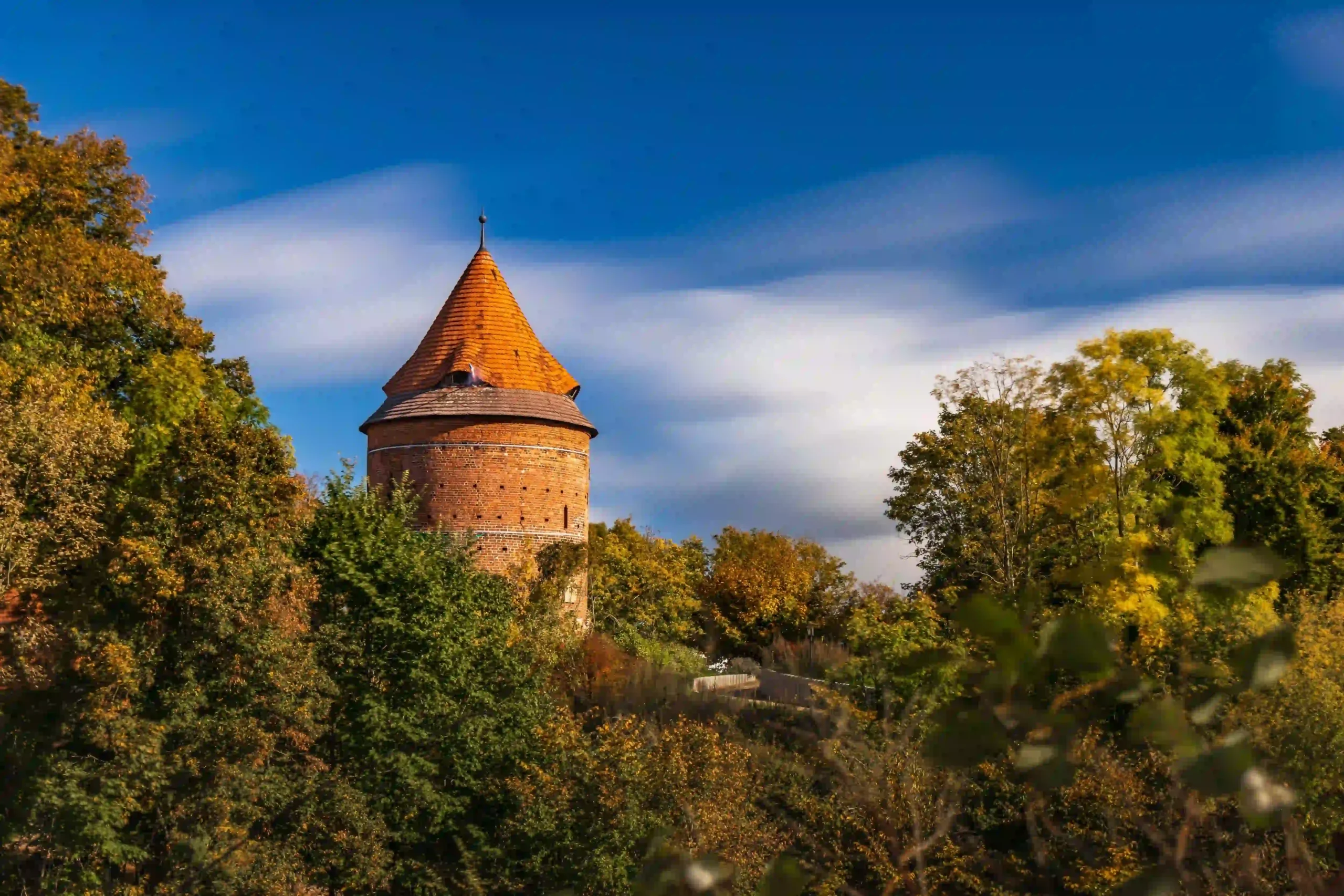 Der Burgturm von Plau am See, ein runder Backsteinturm mit spitzem, orangerotem Kegeldach, umgeben von bunten Herbstbäumen unter einem blauen Himmel mit verschwommenen Wolken.