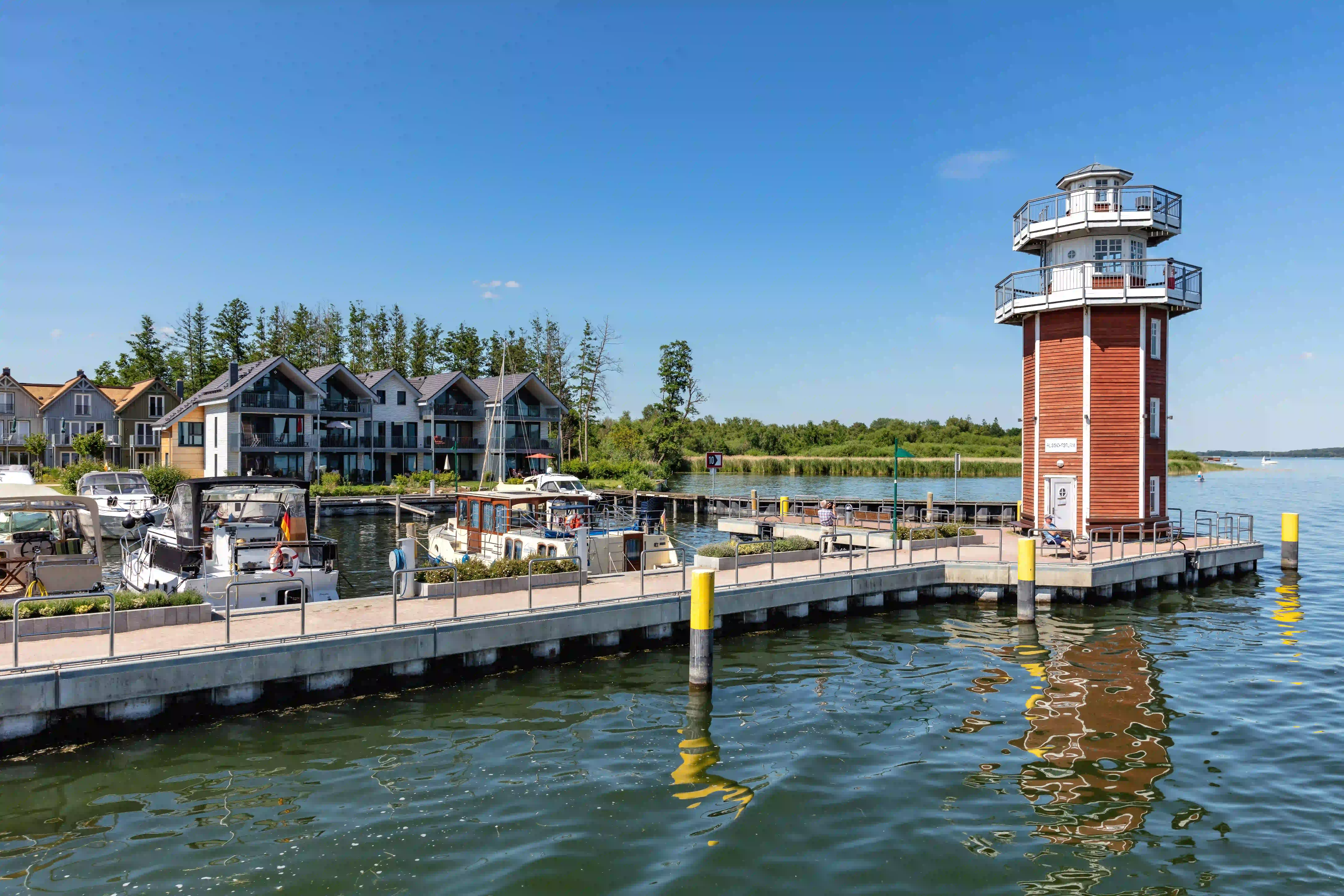 Der Leuchtturm von Plau am See, ein rot-weiß gestreifter Turm am Ende eines langen Holzstegs im See, mit Booten und weiteren Gebäuden am Ufer im Hintergrund unter strahlend blauem Himmel.