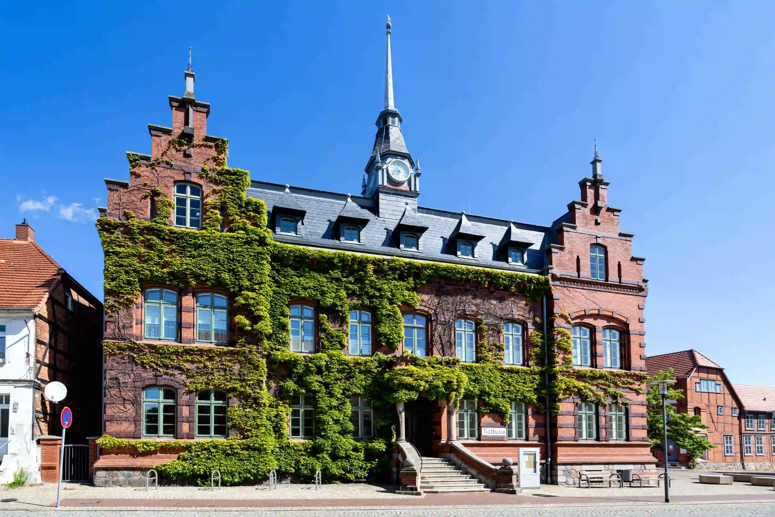 Das Rathaus von Plau am See, ein imposantes Backsteingebäude mit Erkern und einem mittigen Uhrenturm mit spitzem Dach, teilweise von Efeu bewachsen, unter klarem blauem Himmel.