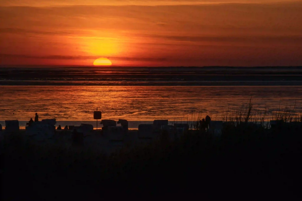 Norddeich mit Hund: Perfekter Nordsee-Urlaub mit Vierbeiner 9 Ein lebhafter Sonnenuntergang in kräftigem Orange über dem Wattenmeer von Norddeich, mit Strandkörben im Vordergrund und der untergehenden Sonne am Horizont. Der perfekte Ausklang eines Tages in Norddeich mit Hund.