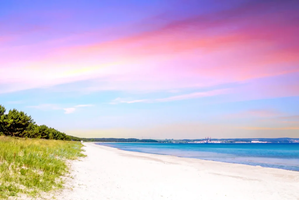 Urlaub mit Hund in Prora auf Rügen – Der perfekte Guide 2 Sonnenuntergang über Prora auf Rügen mit ruhigem Meer, sandigem Strand, grüner Küstenvegetation und fernen Industrieanlagen.