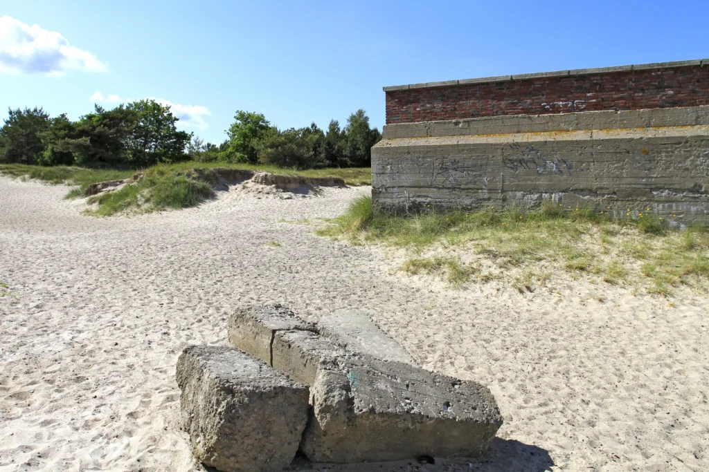 Urlaub mit Hund in Prora auf Rügen – Der perfekte Guide 9 Sonnige Dünenlandschaft mit Betonblöcken und alter Mauer der Prora auf Rügen, umgeben von Sand, Gras und blauem Himmel