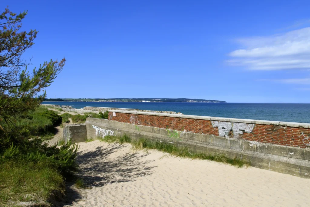 Urlaub mit Hund in Prora auf Rügen – Der perfekte Guide 11 Sonniger Küstenblick mit Strand, Weg und Mauer, im Hintergrund Prora auf Rügen am ruhigen Meerufer.