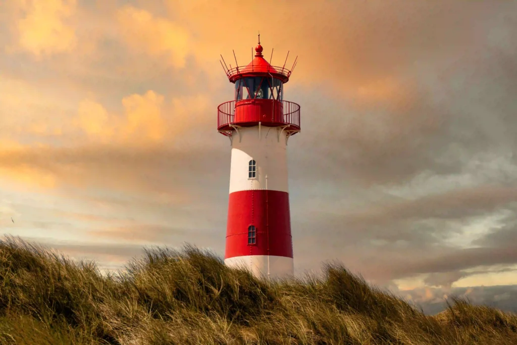 Sylt mit Hund: Dein ultimativer Guide für den perfekten Inselurlaub 10 Rot-weißer Leuchtturm auf einer bewachsenen Düne auf Sylt, umgeben von Gräsern und einem dramatischen Himmel – schöne Ausflugsziele auf Sylt mit Hund.