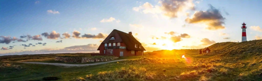 Sylt mit Hund: Dein ultimativer Guide für den perfekten Inselurlaub 2 Idyllisches Reetdachhaus und Leuchtturm auf Sylt bei Sonnenuntergang mit goldenem Licht, das die Landschaft erhellt – ein Traum für den Urlaub auf Sylt mit Hund.