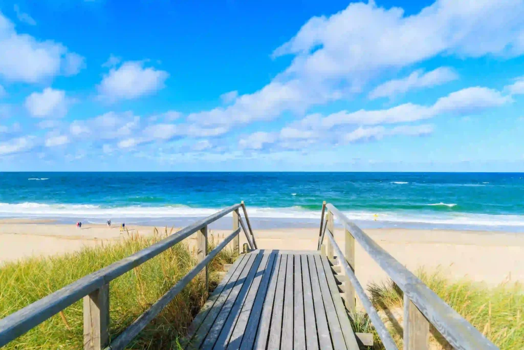 Sylt mit Hund: Dein ultimativer Guide für den perfekten Inselurlaub 5 Holzsteg zum Strand auf Sylt mit weitem Blick aufs Meer und blauem Himmel. Am Horizont sind in der Ferne Spaziergänger zu erkennen – ideal für einen Urlaub auf Sylt mit Hund.