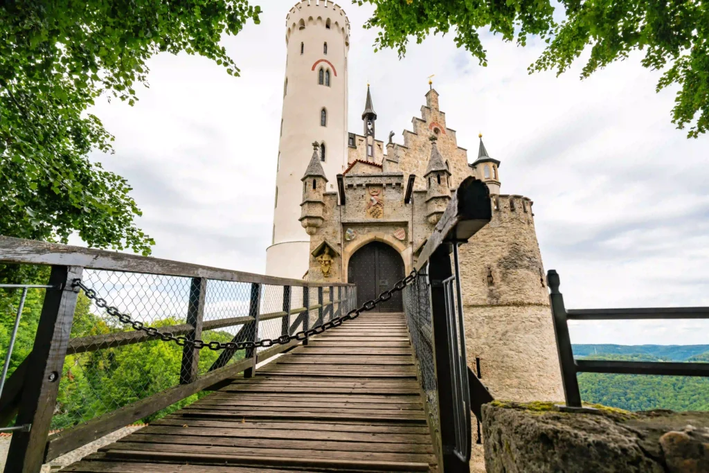 Wandern mit Hund am Schloss Lichtenstein erleben 3 Der Eingang zum Schloss Lichtenstein, mit einer hölzernen Brücke, die zum Haupttor führt, umrahmt von grünen Blättern.