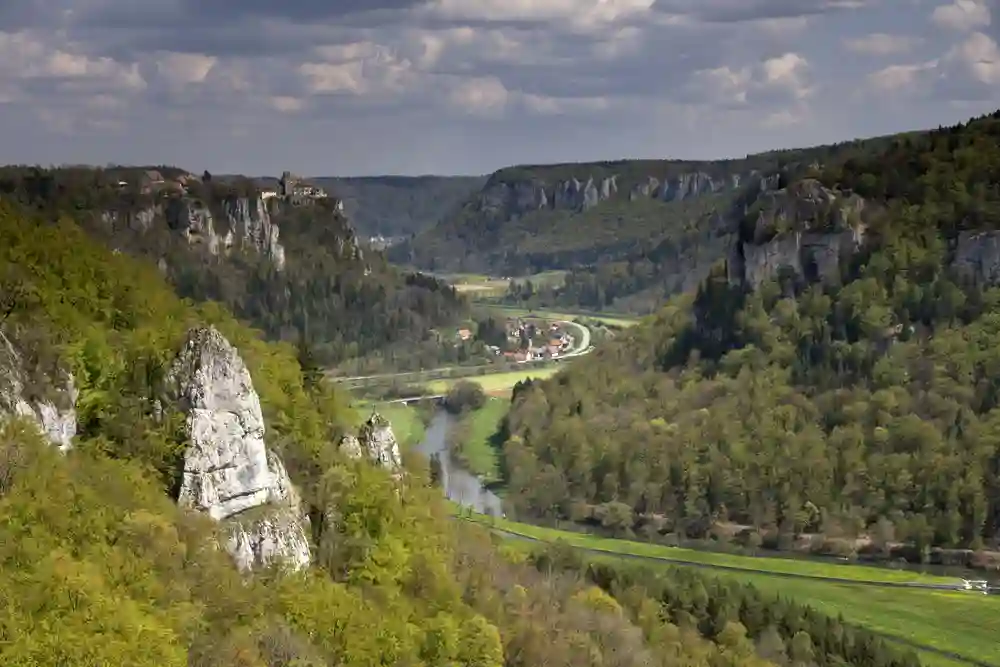 Wandern mit Hund im Oberen Donautal perfekt erleben 3 Eine weite Ansicht des Oberen Donautals mit einem Fluss, der sich durch ein Tal mit bewaldeten Hügeln und markanten Felsformationen windet, und einer kleinen Siedlung in der Ferne.