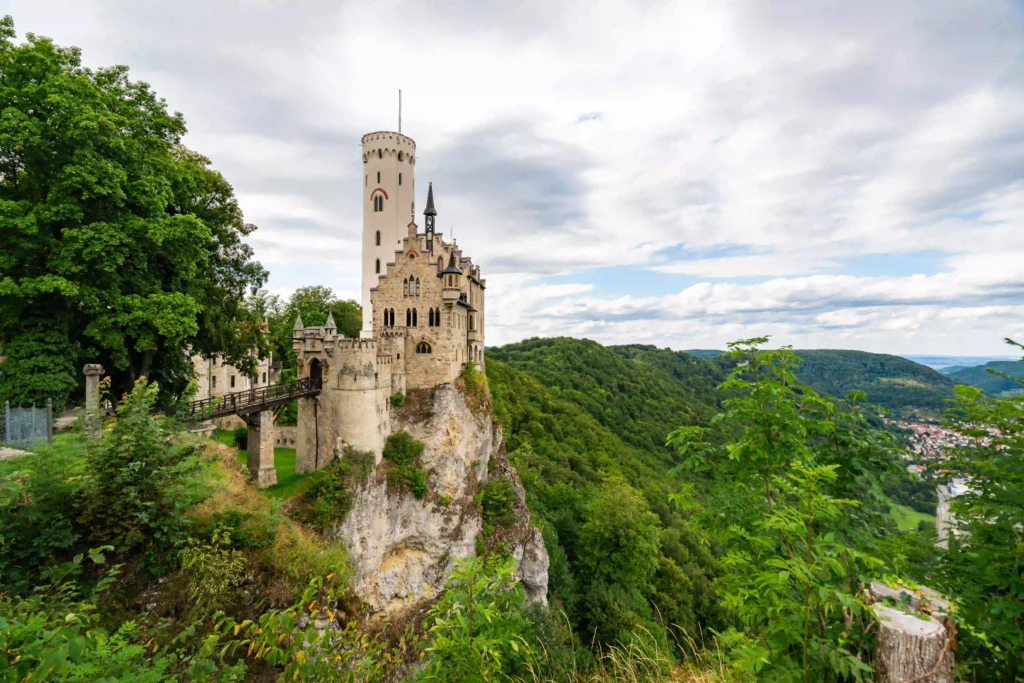Wandern mit Hund am Schloss Lichtenstein erleben 4 Schloss Lichtenstein, eine majestätische Burg, thront auf einem Felsen, umgeben von grünen Bäumen und einem bewölkten Himmel.