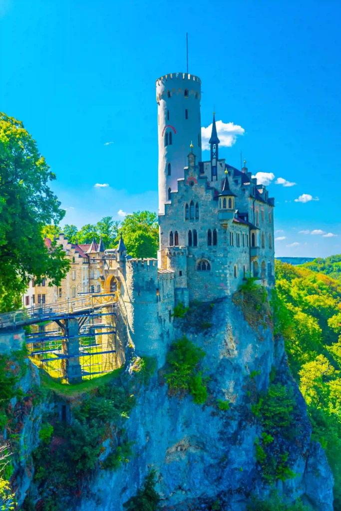 Wandern mit Hund am Schloss Lichtenstein erleben 7 Schloss Lichtenstein in leuchtenden Farben, ein malerischer Anblick, der auf einem Felsen inmitten üppiger grüner Wälder unter strahlend blauem Himmel thront.