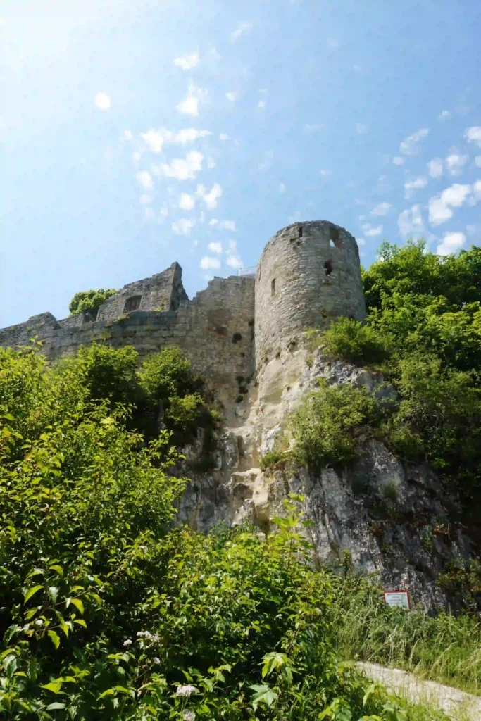 Wandern mit Hund an der Burg Hohenurach erleben 4 Ansicht der Ruine der Burg Hohenurach, von unten nach oben fotografiert, mit einem runden Turm und Mauerresten auf einem Felsvorsprung, umgeben von dichtem Grün unter hellem Himmel.
