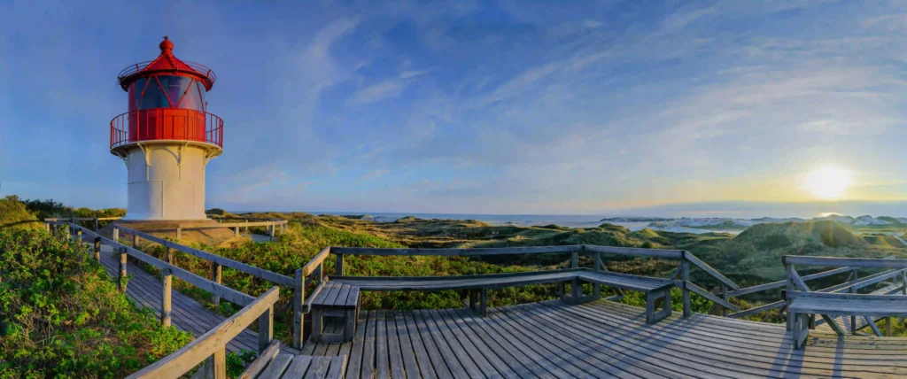 Amrum mit Hund: Perfekter Inselurlaub für Vierbeiner 8 Panoramablick auf den Amrum Leuchtturm bei Sonnenuntergang, umgeben von Dünen und einem Holzsteg, eine schöne Kulisse für Urlaub in Amrum mit Hund.