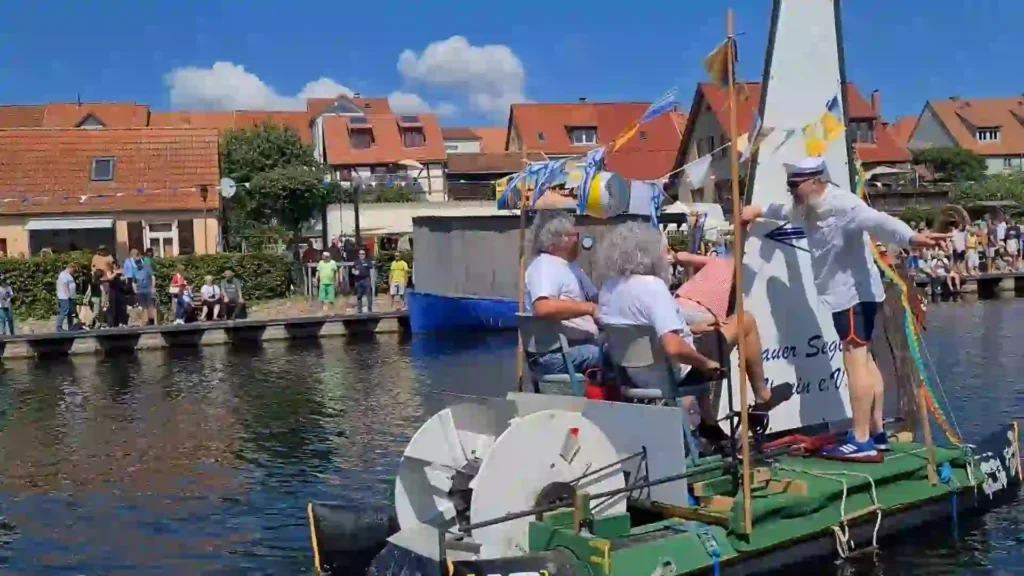 Badewannenrally Plau am See 2025 – Spaß und Action am Wasser 11 Ein Team in weißen Oberteilen navigiert ein grünes, mit Schaufelrädern ausgestattetes Floß bei der Badewannenrally in Plau am See. Zuschauer säumen das Ufer der charmanten Stadtkulisse.
