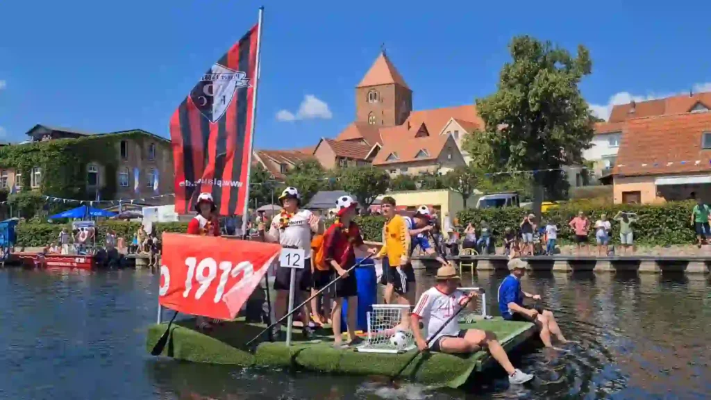 Badewannenrally Plau am See 2025 – Spaß und Action am Wasser 6 Ein Team auf einem grünen Floß mit Fußball-Motiven und einer großen rot-schwarzen Flagge nimmt an der Badewannenrally in Plau am See teil. Im Hintergrund die historische Stadtkulisse mit Kirchturm und jubelnden Zuschauern.