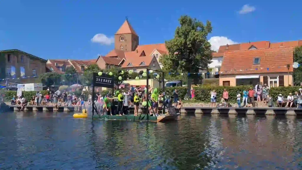 Badewannenrally Plau am See 2025 – Spaß und Action am Wasser 5 Die Startzone der Badewannenrally in Plau am See mit Teilnehmern in leuchtend grüner Kleidung unter einem "395 RAVE"-Banner. Im Hintergrund ein Kirchturm und Zuschauer am Ufer entlang des Wassers.