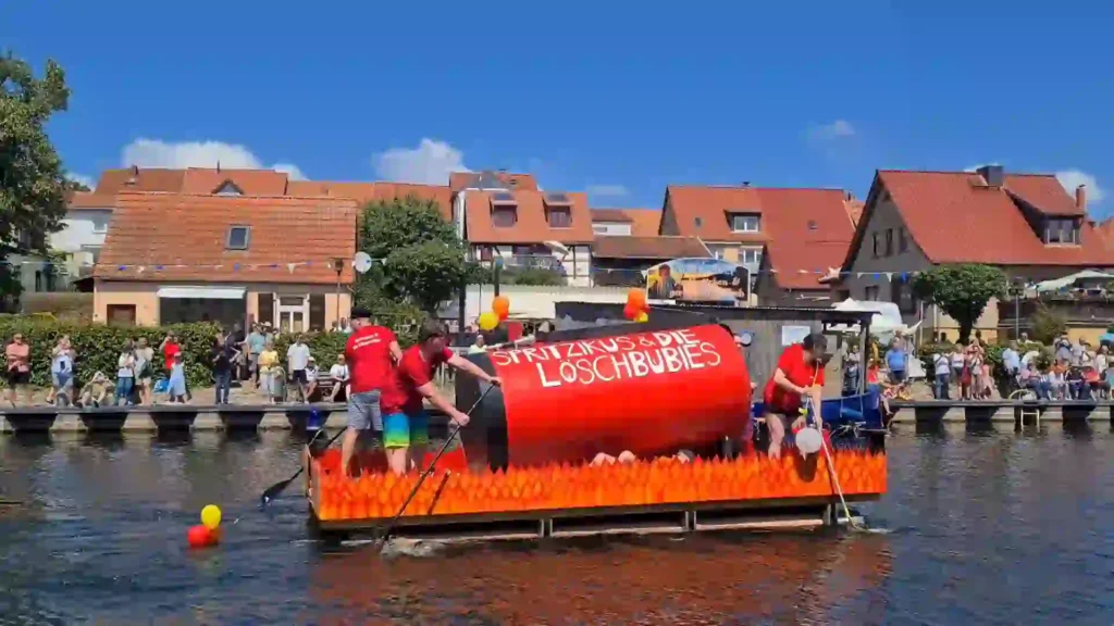 Badewannenrally Plau am See 2025 – Spaß und Action am Wasser 3 Ein rotes, feuerflammenverziertes Floß mit der Aufschrift "SPRITZIKUS & DIE LOSCHBUBIES" und Paddlern fährt bei der Badewannenrally in Plau am See. Viele Zuschauer beobachten das Spektakel vom Ufer aus.
