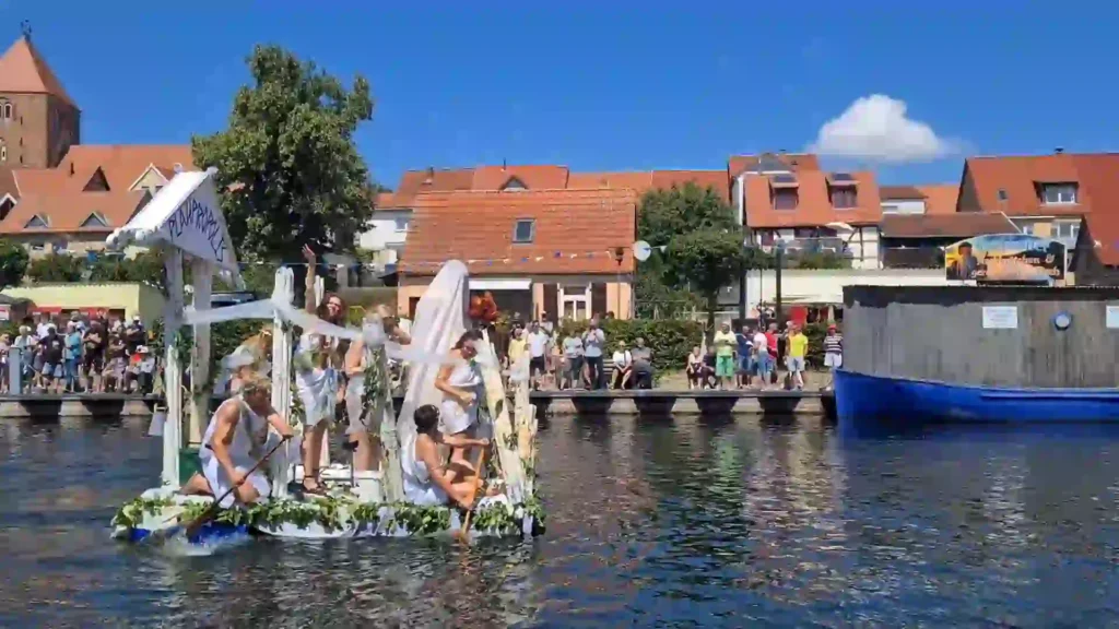 Badewannenrally Plau am See 2025 – Spaß und Action am Wasser 14 Ein aufwändig als griechischer Tempel gestaltetes Floß mit verkleideten Teilnehmern fährt auf dem Wasser der Badewannenrally in Plau am See. Im Hintergrund sind Zuschauer am Ufer und die Häuser der Altstadt zu sehen.