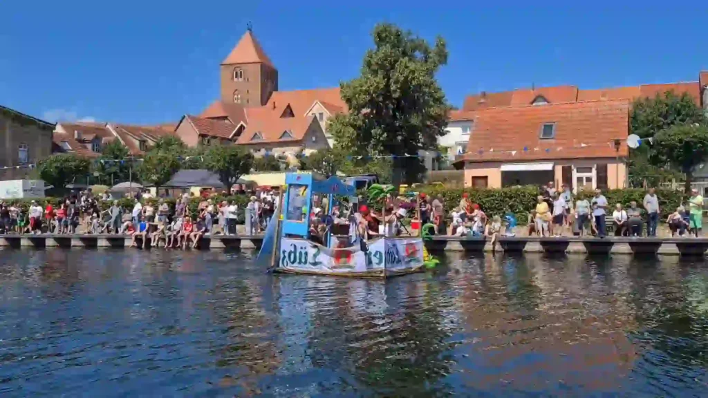 Badewannenrally Plau am See 2025 – Spaß und Action am Wasser 13 Ein buntes, als Kirmes-Bude oder Spielautomat gestaltetes Floß mit mehreren Personen fährt bei der Badewannenrally in Plau am See auf dem Kanal. Am Ufer beobachten zahlreiche Zuschauer das Geschehen vor der Kulisse der historischen Stadt mit Kirchturm.