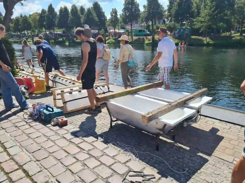 Badewannenrally Plau am See 2025 – Spaß und Action am Wasser 12 Teilnehmer bereiten Badewannen auf einem Steg für die Badewannenrally in Plau am See vor. Werkzeuge und Materialien liegen auf dem Kopfsteinpflaster, im Hintergrund weitere Zuschauer und der Kanal.