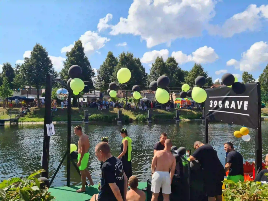 Badewannenrally Plau am See 2025 – Spaß und Action am Wasser 2 Männliche Teilnehmer in Sportkleidung bereiten sich auf der Startplattform der Badewannenrally in Plau am See vor. Im Hintergrund jubelnde Zuschauer am Ufer und gelb-schwarze Ballondekoration unter blauem Himmel.