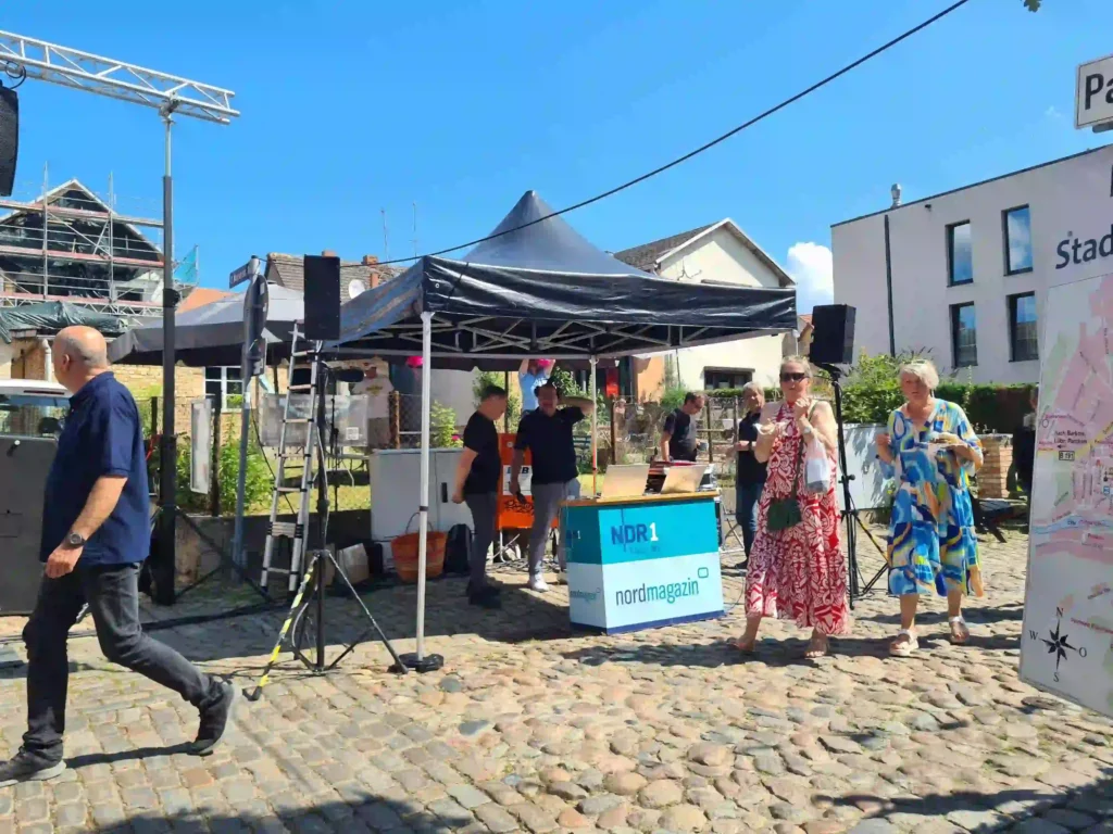 Badewannenrally Plau am See 2025 – Spaß und Action am Wasser 8 Ein NDR-Stand mit "Nordmagazin"-Schild und technischem Equipment auf gepflastertem Boden bei der Badewannenrally in Plau am See. Passanten schlendern vorbei, im Hintergrund Gebäude.