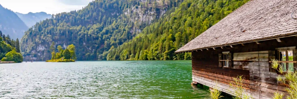 Urlaub am Königssee mit Hund: Tipps & Highlights 9 Königssee mit Hund: Ruhiger See mit bewaldeten Bergen, Holzhaus am Ufer und kleinen Booten in malerischer Naturkulisse.