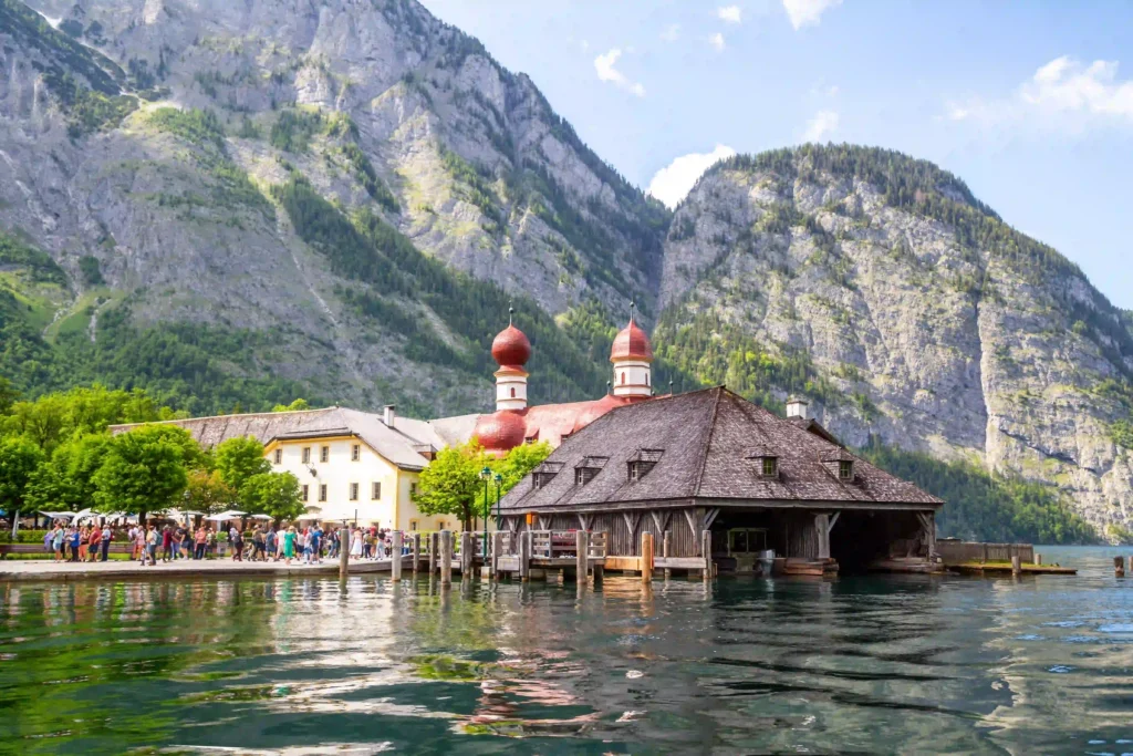 Urlaub am Königssee mit Hund: Tipps & Highlights 11 Königssee mit Hund: Blick auf ruhigen See, St. Bartholomä, Promenade und Alpenberge bei sonnigem Himmel.