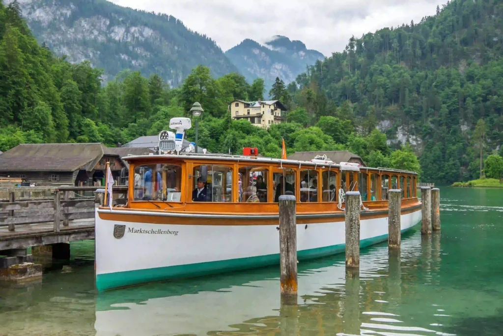 Urlaub am Königssee mit Hund: Tipps & Highlights 10 Passagierschiff auf klarem grünen Königssee mit Hund, umgeben von Bergen, Wald und ruhigem Wasser bei bewölktem Himmel