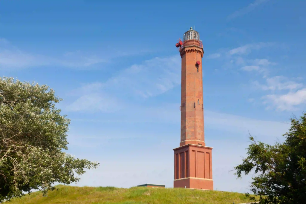 Norderney mit Hund: Top Wanderungen & Ausflugsziele an der Nordsee 2 Der imposante rote Leuchtturm von Norderney ragt majestätisch in den blauen Himmel empor, umgeben von grünen Hügeln und Bäumen – ein Wahrzeichen, das man mit Hund erkunden kann.