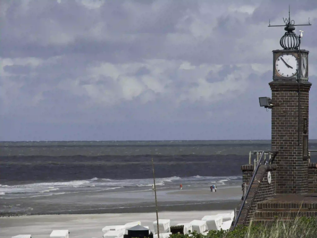 Urlaub auf Wangerooge mit Hund: Dein Guide für perfekte Ferien mit Vierbeiner 8 Wangerooge Strand mit dem historischen Uhrturm und der rauen Nordsee, im Hintergrund Strandkörbe und Spaziergänger mit Hunden.