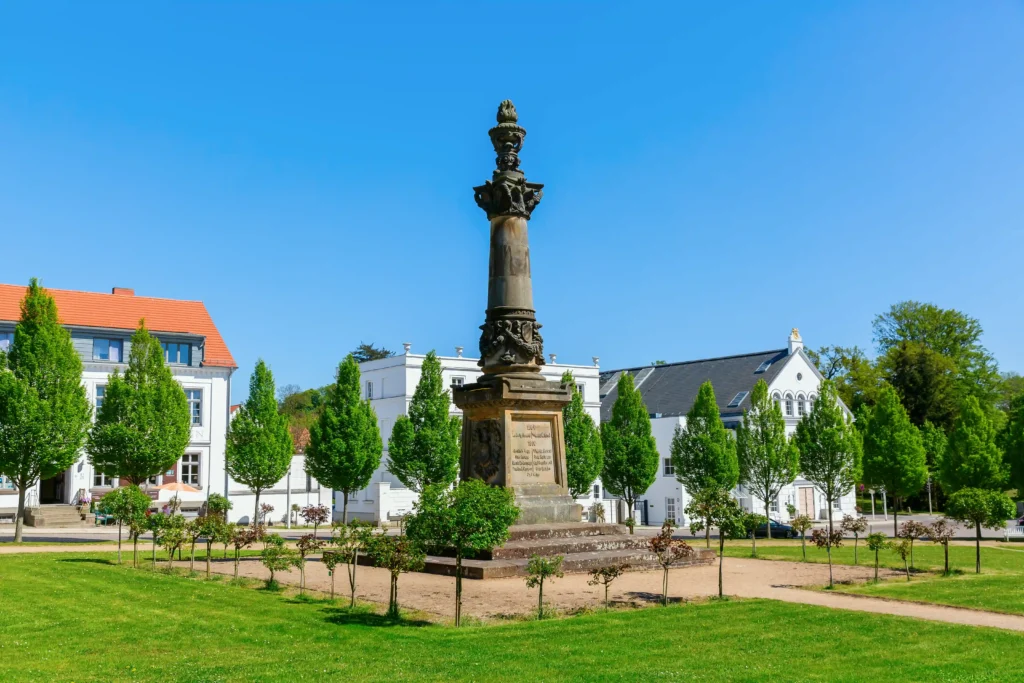 Urlaub in Putbus mit Hund – Der perfekte Guide für Rügen 9 Steinernes Denkmal im sonnigen Park von Putbus mit grüner Wiese, Blumen und historischen Gebäuden im Hintergrund