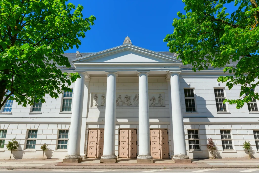 Urlaub in Putbus mit Hund – Der perfekte Guide für Rügen 3 Neoklassizistisches weißes Gebäude mit Portikus und Säulen in Putbus, umrahmt von grünen Bäumen bei blauem Himmel.