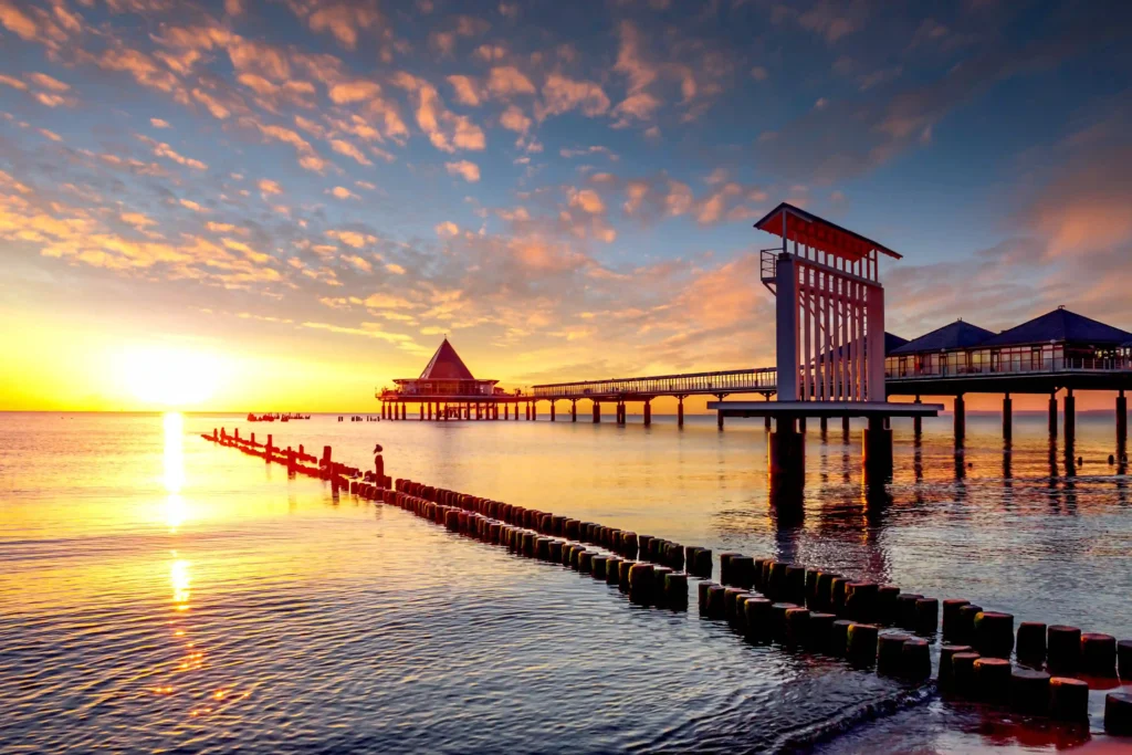 Urlaub mit Hund in Heringsdorf: Dein perfekter Ostsee-Guide 24 Sonnenuntergang über Heringsdorf mit Hund am Pier, ruhiges Meer, bunte Himmel und Möwen auf Holzpfählen am Ufer.