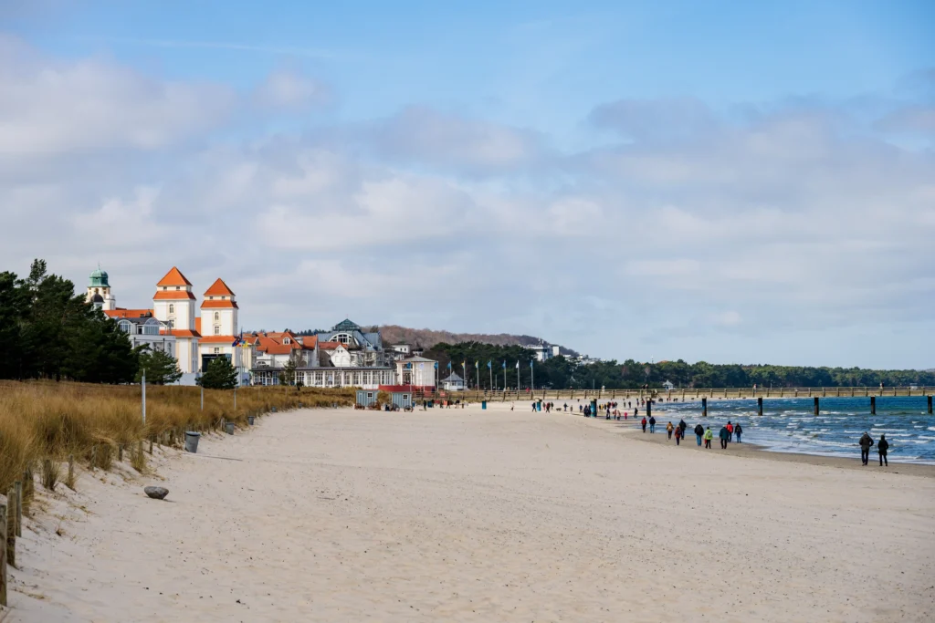 Dein perfekter Ausflug zum Hundestrand Binz 7 Strandpanorama von Binz mit Sand, Menschen, Pier, Gebäuden mit roten Dächern und bewölktem Himmel im Hintergrund