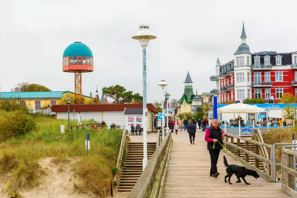 Urlaub mit Hund in Zinnowitz: Dein perfekter Ostseeurlaub 2 Zinnowitz mit Hund: Person mit schwarzem Hund auf grünem Leine spaziert über Holzpromenade am Strand bei bewölktem Himmel.